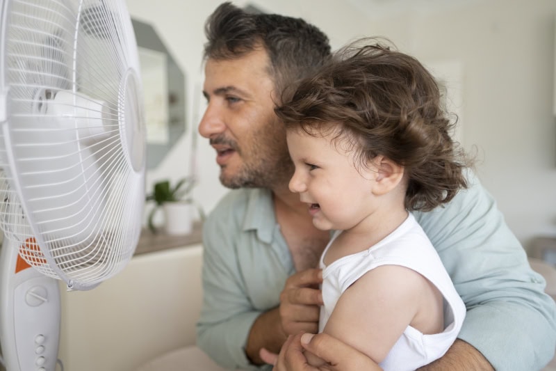 Father holding son up to fan, trying to cool off due to their home's AC problems.