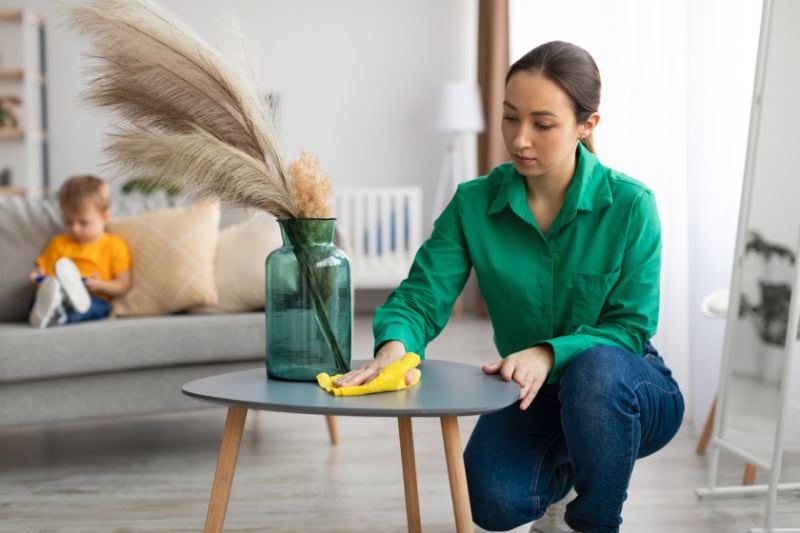 Mother cleaning off table in living room to reduce dust in her Charlotte, North Carolina, home. Son playing game on couch.